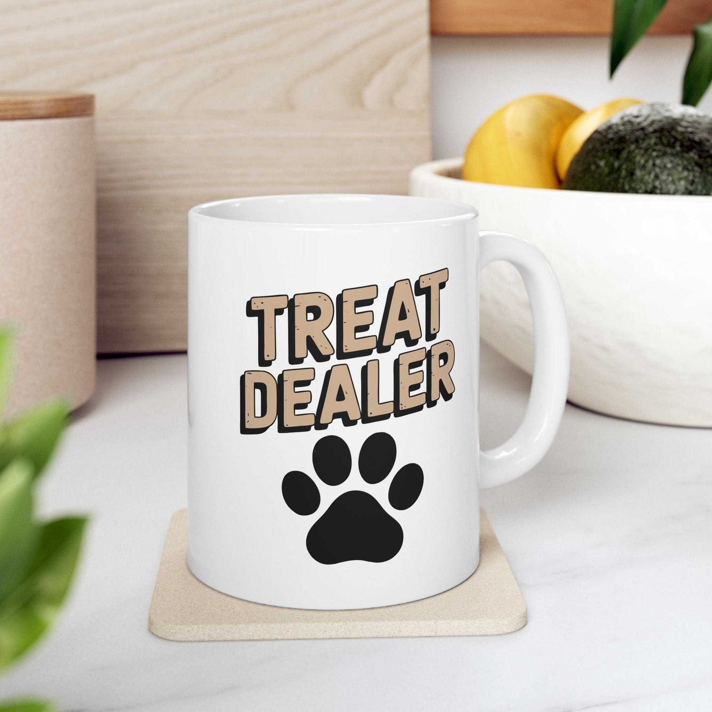 White mug with 'Treat Dealer' text and paw print on a coaster, placed on a surface with plants and containers in the background.