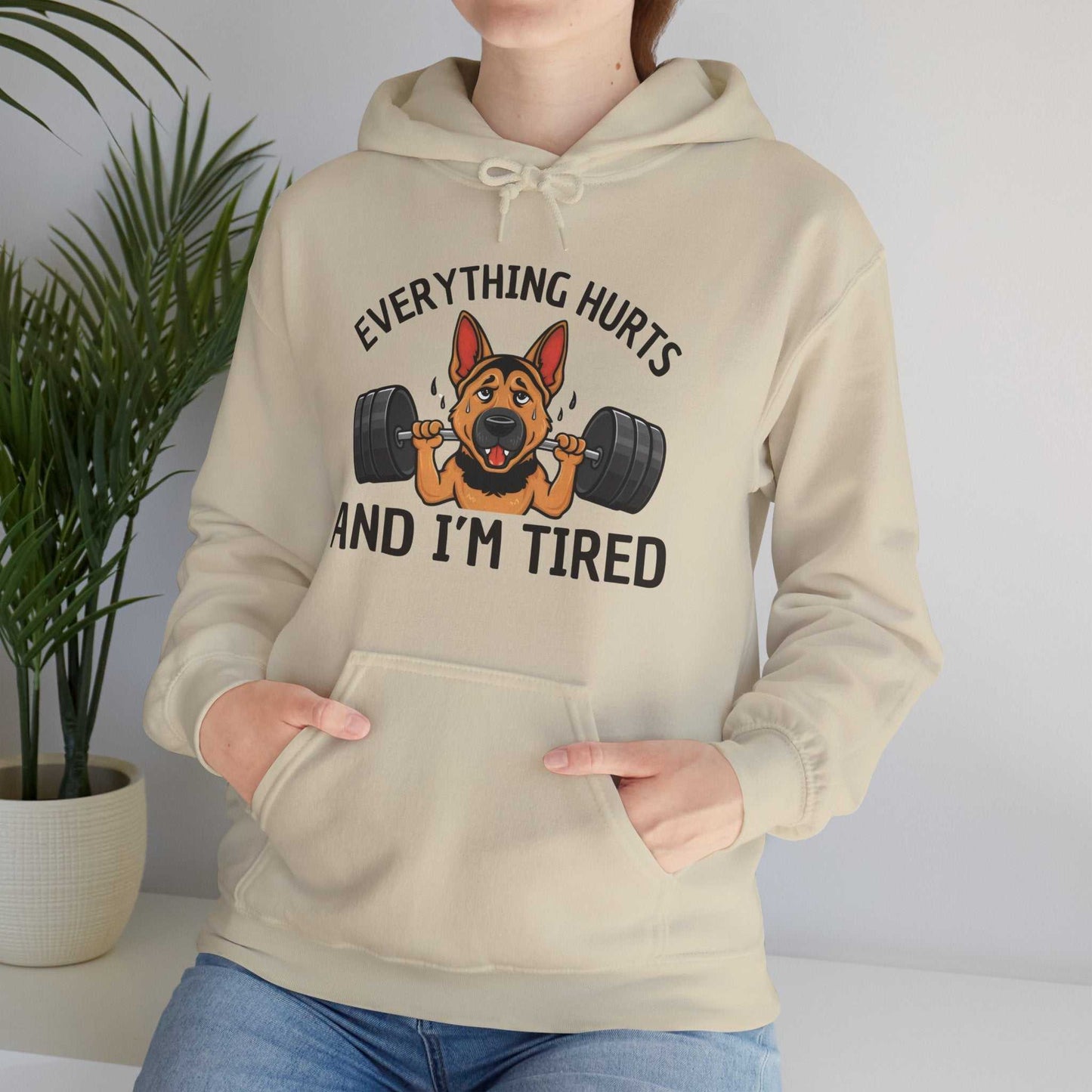 Person wearing a beige hoodie with a graphic of a dog lifting weights and text 'Everything hurts and I'm tired'.