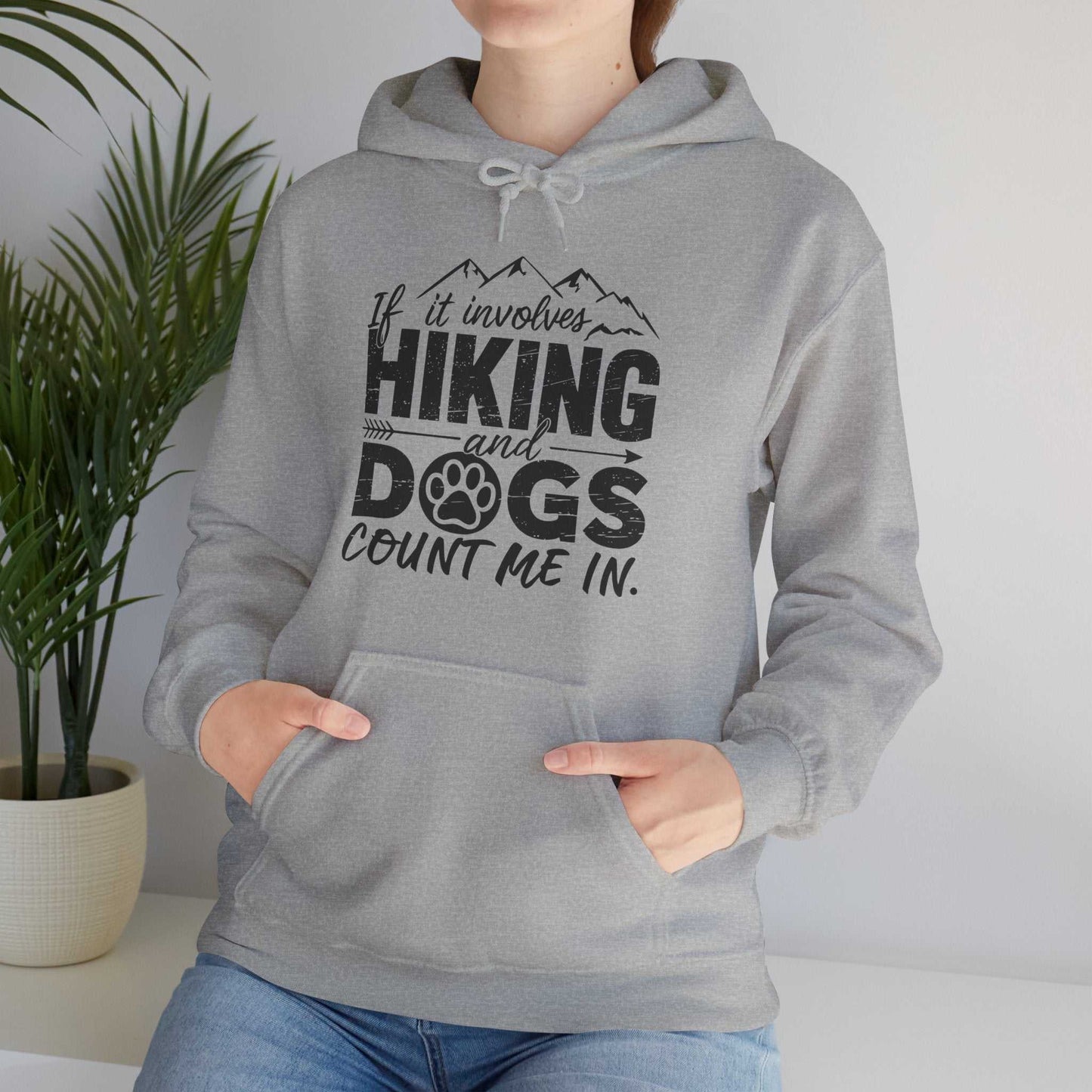 Person wearing a gray hoodie with a hiking and dogs design, standing indoors with a plant in the background.