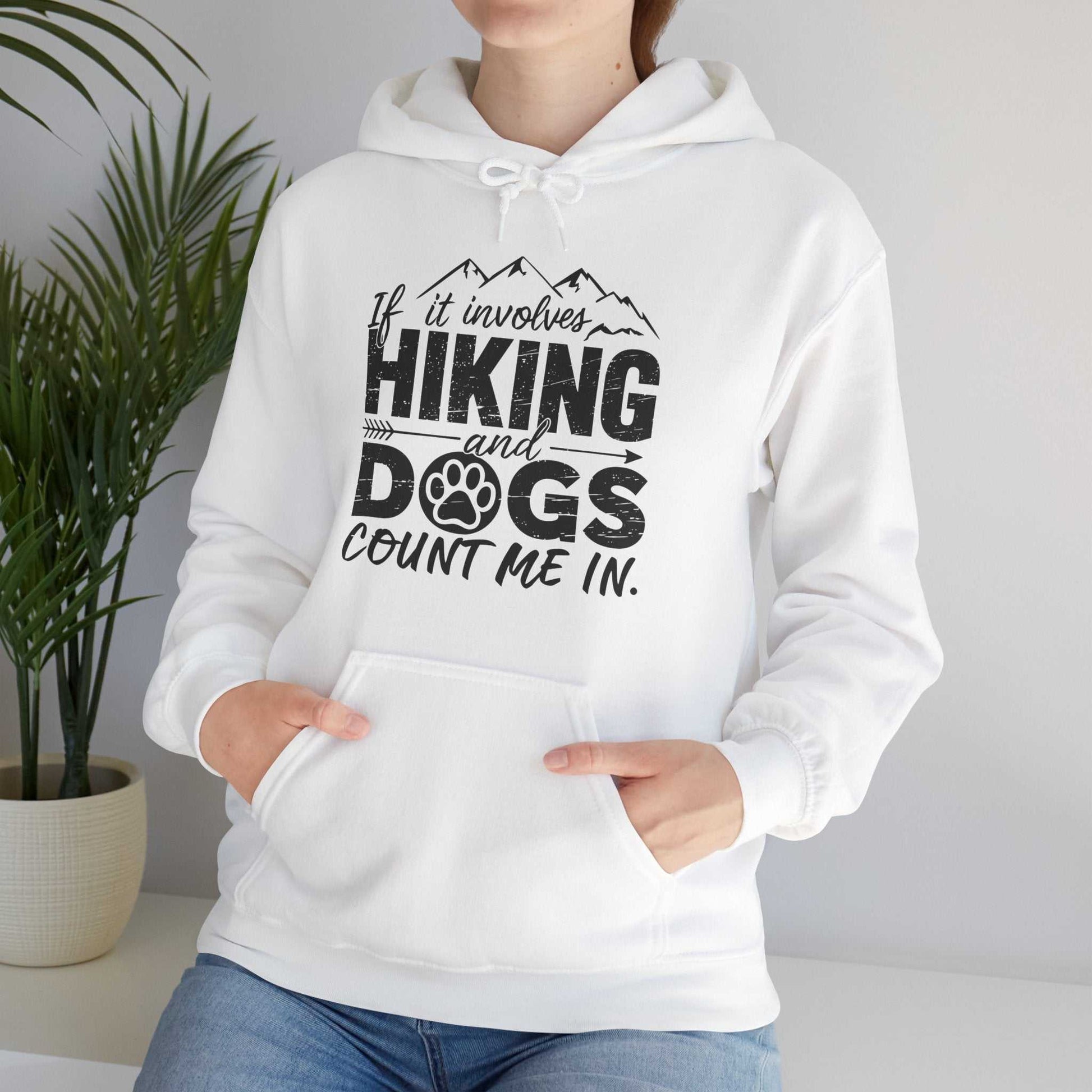 White hoodie with black text 'If it involves hiking and dogs count me in.' worn by a person, with a plant in the background.