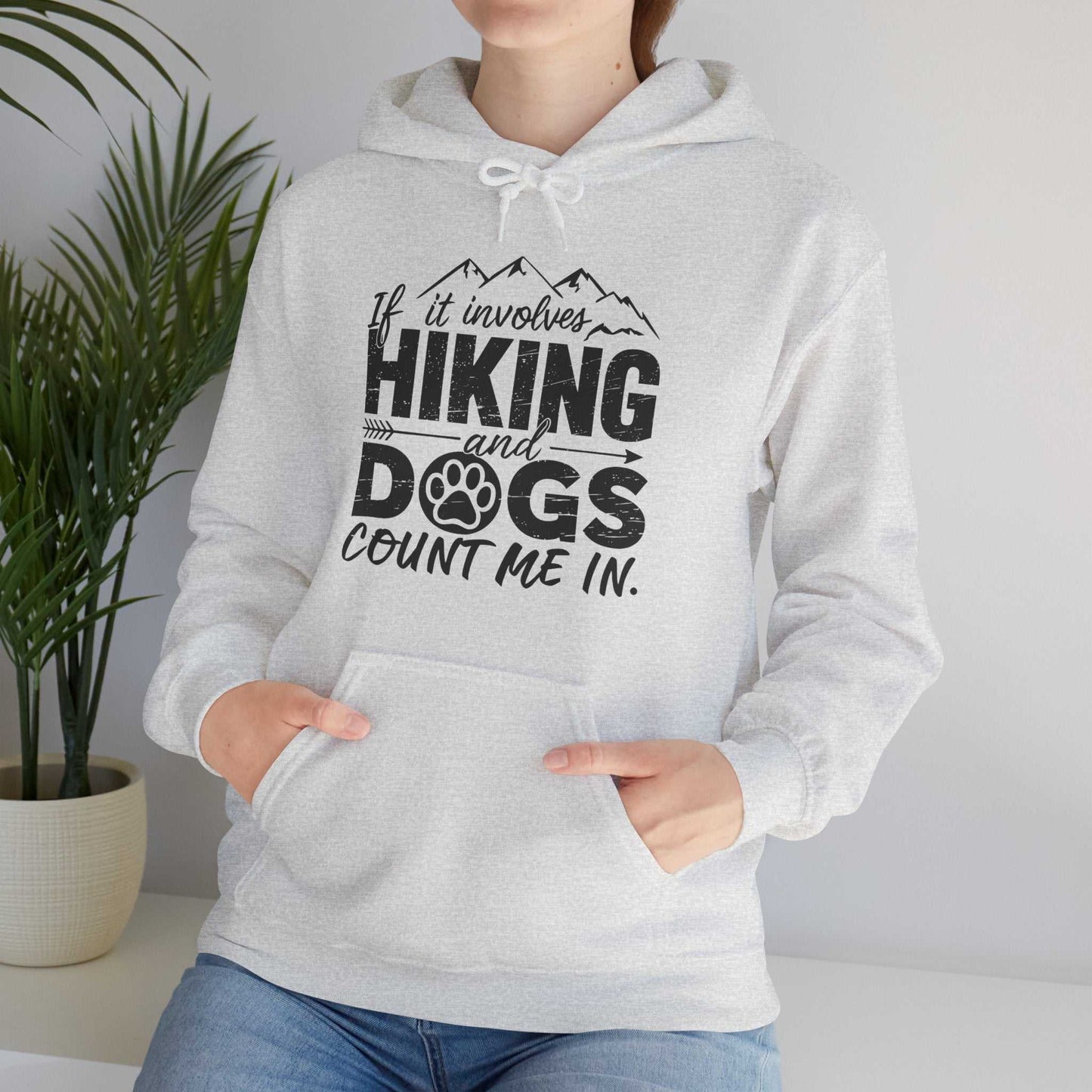 Person wearing a gray hoodie with a hiking and dogs design, standing indoors next to a plant.